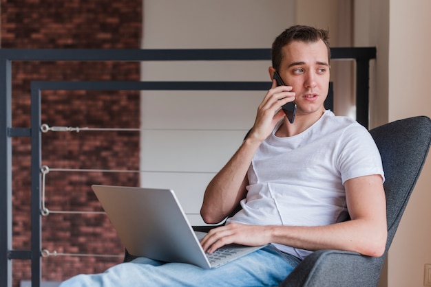 Homem concentrado falando no celular e sentado na cadeira com laptop Foto gratuita