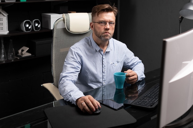 Homem de negócios com óculos, trabalhando no escritório na mesa do computador e tomando café da xícara brilhante Foto Premium