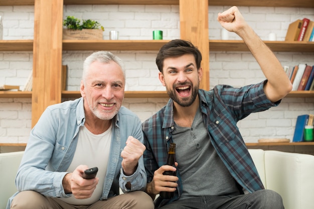 Homem envelhecido com controle remoto e cara chorando jovem com garrafa assistindo tv no sofá Foto gratuita