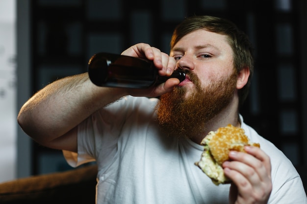 Homem gordo assiste tv, come hambúrguer e bebe cerveja Foto gratuita