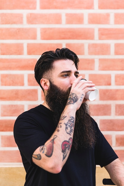 Homem jovem, tendo, tatuagem, ligado, seu, mão, café bebendo, contra, parede tijolo Foto gratuita
