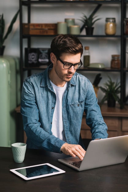 Homem jovem, usando computador portátil, com, xícara café, e, tablete digital, ligado, contador cozinha Foto gratuita