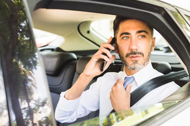 Homem moderno, sentado no carro, ajustando a gravata do pescoço, falando no celular Foto gratuita