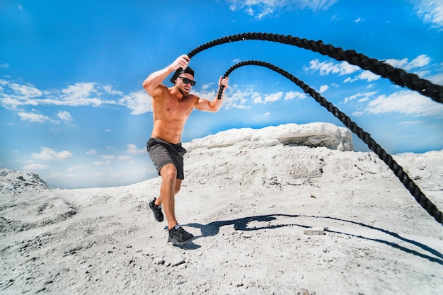 Homem musculoso malhando com cordas de batalha. atlético jovem malhando com cordas de batalha ao ar livre. exercício em forma de esporte. Foto Premium