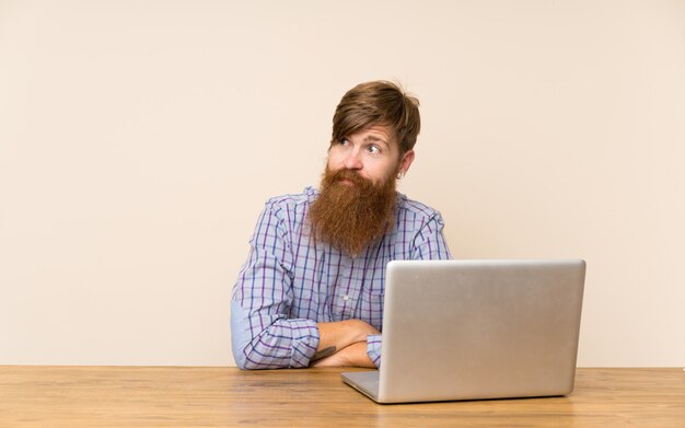 Homem ruivo com barba longa em uma mesa com um laptop em pé e olhando para o lado Foto Premium