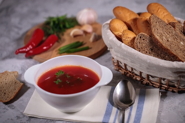 Jantar saboroso e saudável. um prato com sopa de beterraba na mesa, ao lado do tabuleiro, é salsa, endro, cebola verde, alho, pimenta e uma cesta com pão. Foto Premium