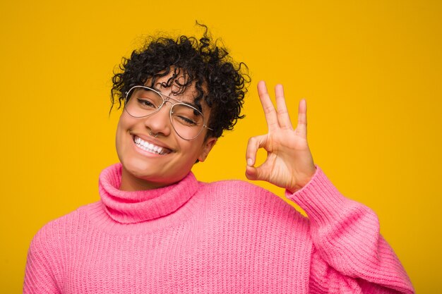 Jovem americana africano vestindo um suéter rosa pisca um olho e mantém um gesto bem com a mão. Foto Premium