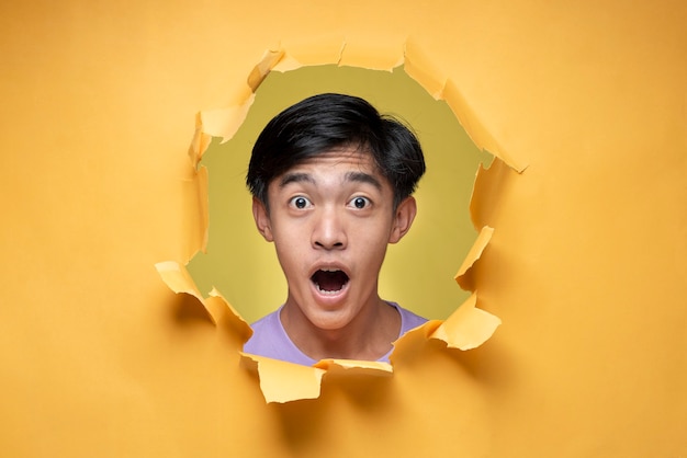 Jovem asiático com expressão chocada e espantada, adolescente, posa através  de um buraco de papel amarelo