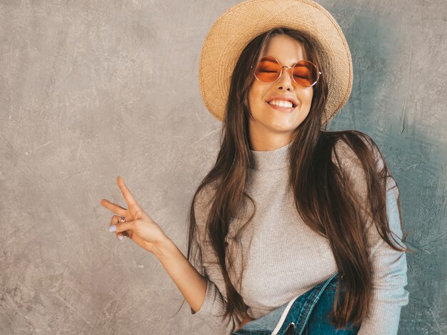 Jovem bela mulher sorridente olhando. menina na moda em roupas de verão casual macacão e chapéu. mostra sinal de paz Foto gratuita