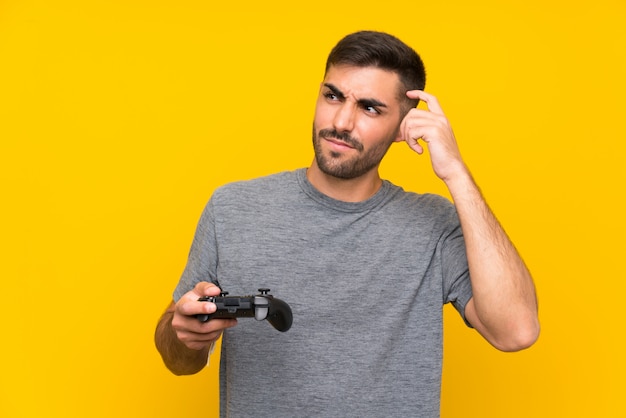 Jovem bonito brincando com um controlador de videogame sobre parede amarela isolada, tendo dúvidas e com a expressão do rosto confuso Foto Premium