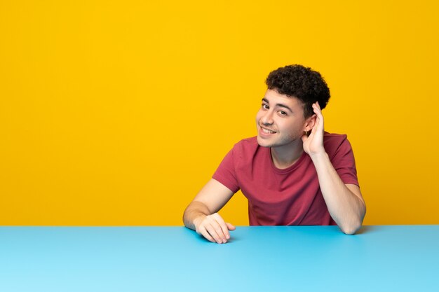 Jovem com parede colorida e mesa ouvindo algo, colocando a mão sobre a orelha Foto Premium