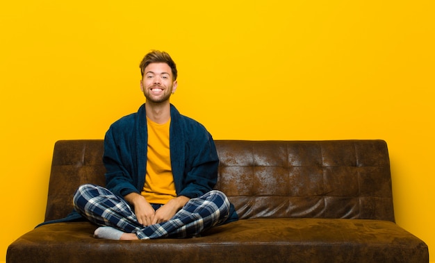 Jovem de pijama, olhando feliz e pateta com um sorriso largo, divertido e maluco e olhos bem abertos. sentado em um sofá Foto Premium