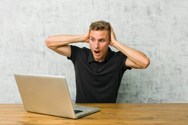 Jovem empreendedor trabalhando com seu laptop em uma mesa cobrindo os ouvidos com as mãos tentando não ouvir som muito alto Foto Premium