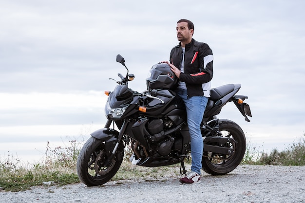 Jovem motociclista com sua moto preta pronta para dirigir, em frente ao mar Foto Premium