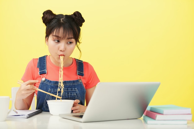 Jovem mulher asiática sentada na mesa, comendo macarrão com pauzinhos e olhando para laptop Foto gratuita
