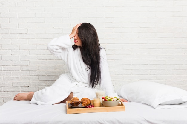 Jovem mulher curvilínea tomando um café da manhã na cama esquecendo algo, batendo na testa com a palma da mão e fechando os olhos. Foto Premium