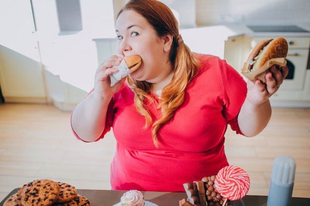 Jovem mulher gorda na cozinha, sentado e comendo comida lixo. estilo de vida não saudável. pedaço de hambúrguer e olhe para a câmera. corpo positivo. doces na mesa. | Foto Premium