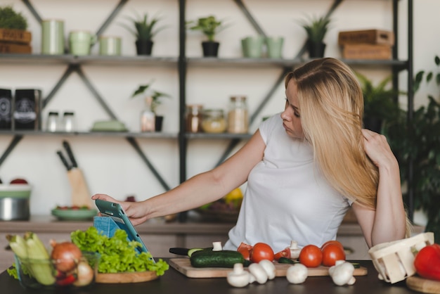 Jovem mulher navegando na tabuleta digital com muitos vegetais no balcão da cozinha Foto gratuita