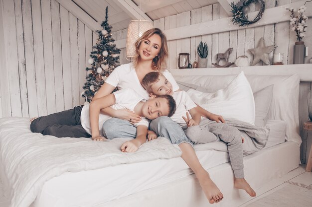 Linda mãe e seus dois filhos, abraçando no quarto branco estilo escandinavo, com árvore de natal Foto Premium