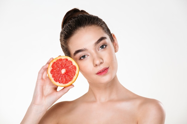 Lindo retrato de mulher bonita seminua com maquiagem natural, segurando a toranja suculenta perto do rosto e olhando Foto gratuita
