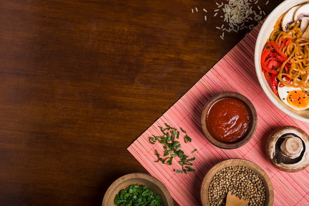 Macarrão ramen em estilo asiático com molhos; cebolinha e sementes de coentro no placemat sobre a mesa de madeira Foto gratuita