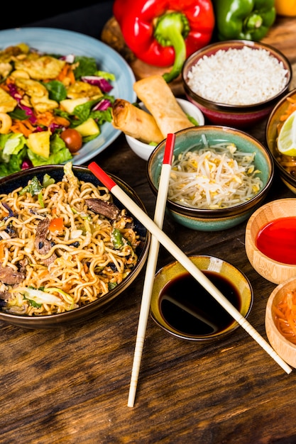 Macarrão tailandês; salada; rolinho primavera; arroz; brotos de feijão; molhos com pauzinhos na mesa de madeira Foto gratuita
