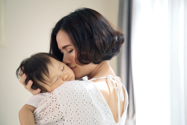 Mãe bonita asiática que abraça o bebê de sono em seus braços e que beija a criança delicadamente. a mãe fecha os olhos enquanto segura a cabeça do bebê para descansar no ombro. toque de amor e relacionamento familiar. Foto Premium
