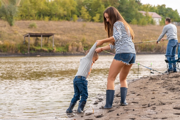 Mãe com um filho pequeno caminha ao longo da costa arenosa do lago em botas de borracha. sair com as crianças na natureza, longe da cidade Foto Premium