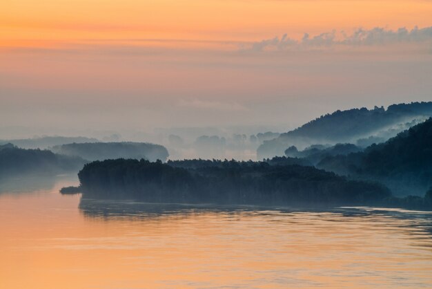 Manhã neblina mística acima amplo vale do rio. brilho de ouro do amanhecer no céu. margem do rio com a floresta sob o nevoeiro. luz solar refletida na água ao nascer do sol. paisagem atmosférica colorida de natureza majestosa. Foto Premium