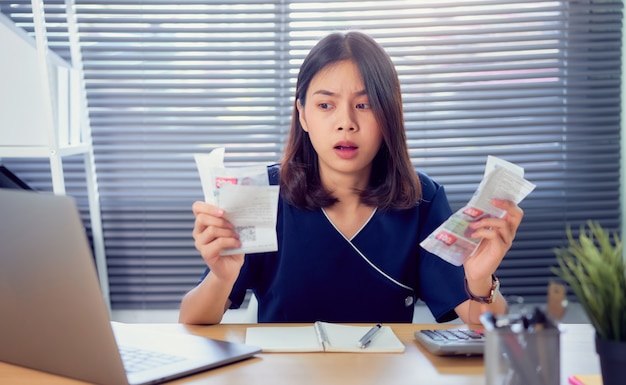 Mão de mulher asiática de rosto chocado segurando a conta de despesa e cálculo mensal sobre as contas da dívida à mesa no escritório em casa. Foto Premium