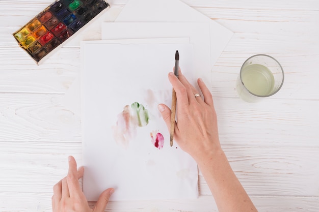 Mãos de mulher com pincel perto de papel com borrões, vidro e conjunto de cores de água Foto gratuita