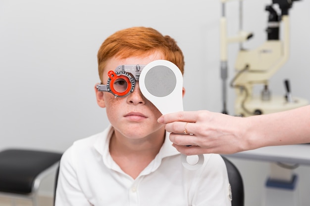 Médica, verificando a visão do garoto sardento com quadro de julgamento optometrista na clínica Foto gratuita