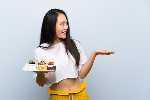 Menina asiática de adolescente segurando muitos mini bolos diferentes com expressão facial de surpresa Foto Premium