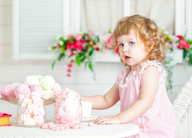 Menina curly bonito pequena em um vestido cor-de-rosa com laço e às bolinhas que sentam-se na tabela e que comem doces diferentes. Foto Premium