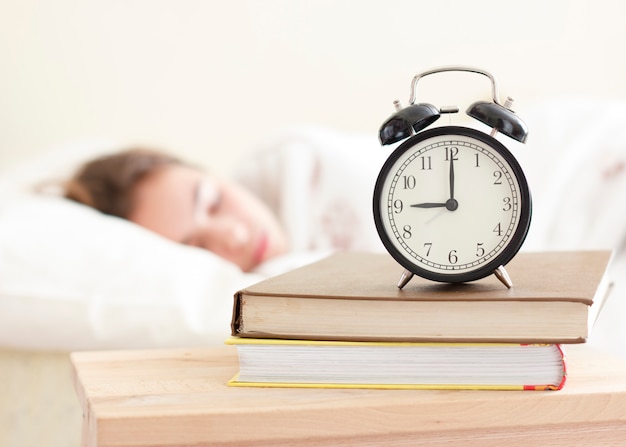 Menina do adolescente que dorme em uma cama branca. despertador em primeiro plano em uma pilha de livros Foto Premium