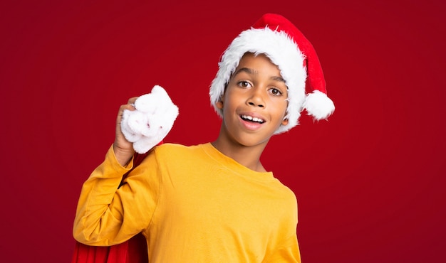 Menino afro-americano com chapéu de natal e levar uma sacola com presentes e fazendo o gesto de surpresa sobre fundo vermelho Foto Premium