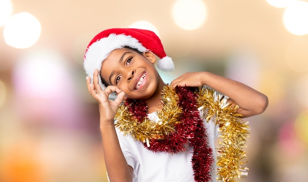 Menino afro-americano com chapéu de natal, mostrando sinal de ok com os dedos sobre fundo desfocado Foto Premium