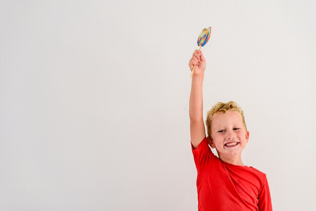 Menino com a camisa vermelha no fundo branco que come um divertimento colorido e um riso do pirulito. Foto Premium