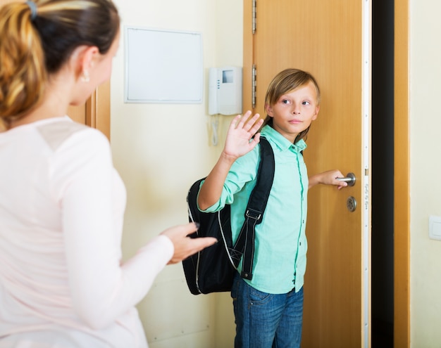 Menino de camisa verde, calça jeans e mochila se despedindo da mãe para ir à escola.