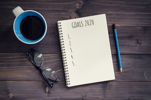 Metas para o ano novo, resolução ou plano de ação 2020. mesa de madeira de escritório com o bloco de notas em branco, lápis, óculos, telefone e xícara de café. Foto Premium