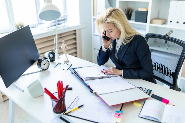 Moça que senta-se na mesa no escritório, falando no telefone e olhando originais. Foto Premium
