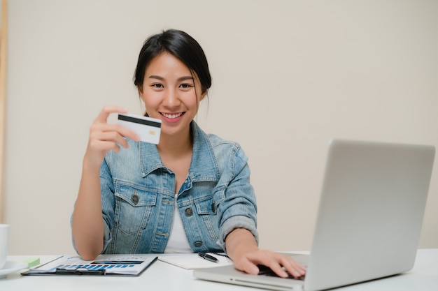 Mulher asiática do negócio esperto bonito que usa o computador ou o portátil que compram a compra em linha pelo cartão de crédito quando vestir o assento ocasional esperto na mesa na sala de visitas em casa. Foto gratuita