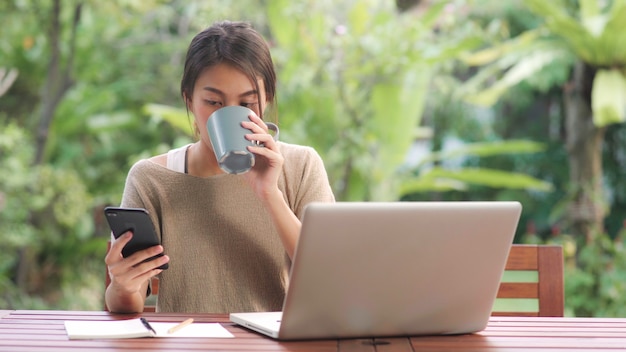 Mulher asiática freelance trabalhando em casa, negócios fêmea trabalhando no laptop e usando telefone celular, bebendo café sentado na mesa no jardim de manhã. Foto gratuita