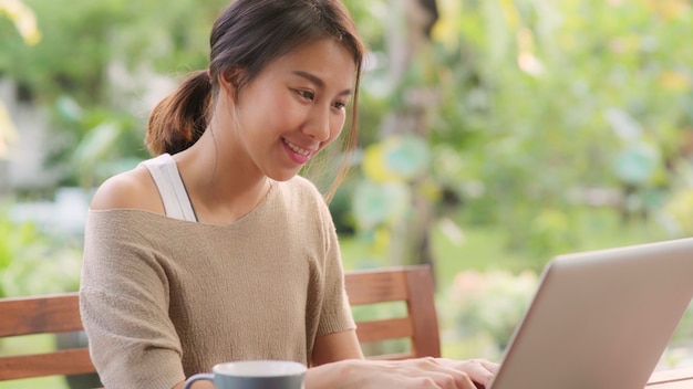 Mulher asiática freelance trabalhando em casa, negócios feminino trabalhando no laptop sentado na mesa no jardim de manhã. mulheres do estilo de vida que trabalham em casa o conceito. Foto gratuita