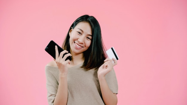 Mulher asiática nova que usa o smartphone que compra a compra em linha pelo cartão de crédito que sente o sorriso feliz na roupa ocasional sobre o tiro cor-de-rosa do estúdio do fundo. Foto gratuita