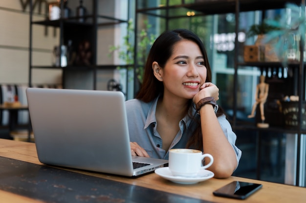 Mulher asiática que trabalha e bebe café no café com sorriso de computador portátil e trabalho feliz Foto Premium