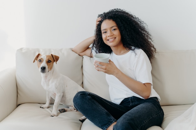 Mulher bonita com cabelo afro, bebe café, posa na sala de estar no sofá com cachorro de raça Foto Premium