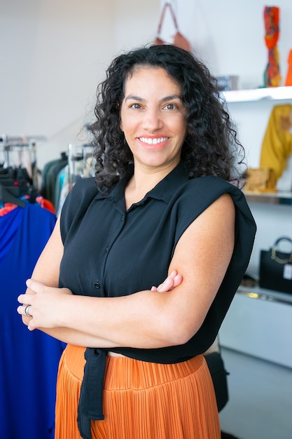 Mulher de cabelos negros latina alegre em pé com os braços cruzados perto do rack com vestidos na loja de roupas, olhando para a câmera e sorrindo. cliente de boutique ou conceito de assistente de loja Foto gratuita