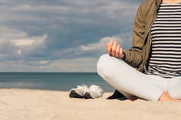 Mulher em uma jaqueta verde e jeans sentado com os pés descalços na praia e meditando Foto Premium