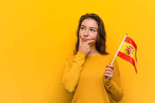 Mulher européia nova que prende uma bandeira espanhola que olha lateralmente com expressão duvidosa e cética. Foto Premium